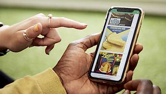 A close up of a hand holding a mobile phone.. On the mobile phone is a quiz with three pictures of desserts including cheesecake and madeira cake. There is another hand to the left with rings on pointing at the phone.