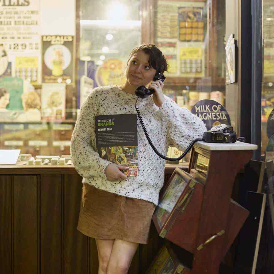 A woman in a white jumper and brown skirt stands holding a rotary telephone receiver to her ear. The telephone is an interactive part of the Museum collection that she is standing in