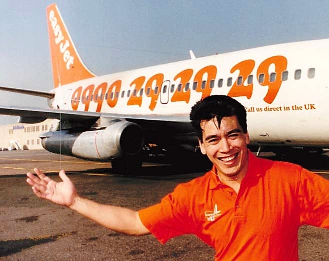 A smiling man in an orange shirt with his arms out, standing in front of a white and orange easyJet plane