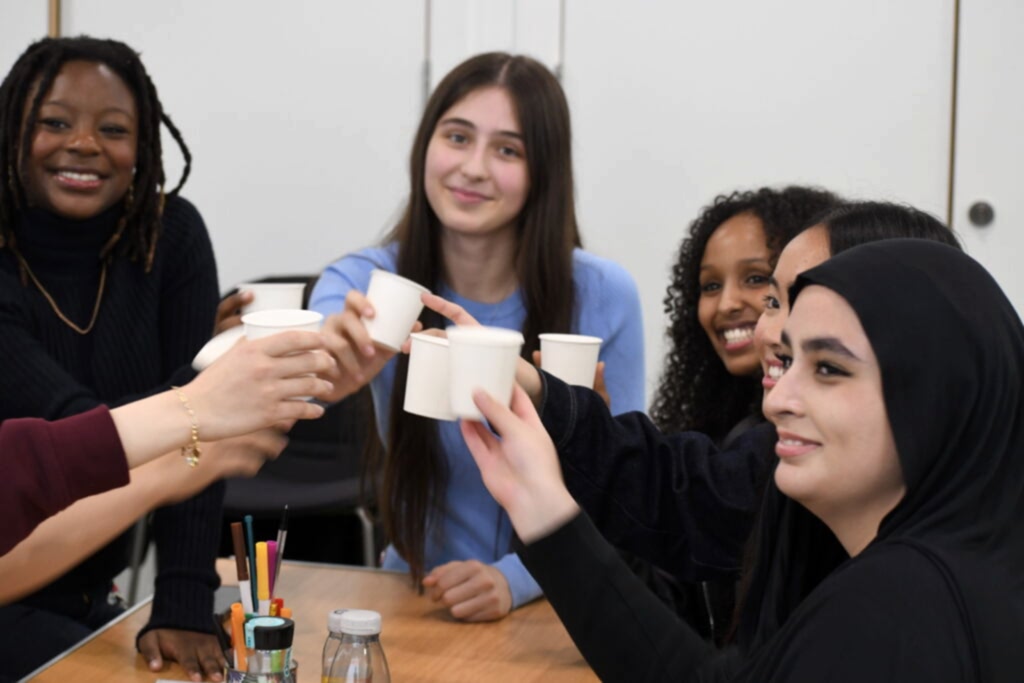 Five school students sit around a table smiling and toasting with cups of smoothie. 